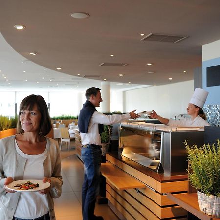 Hotel Neptun Rostock Eksteriør bilde The photo depicts a modern restaurant interior. In the foreground, a woman holding a plate of food looks contemplatively at something in the distance. In the background, a man is interacting with a chef, who is wearing a white chef's hat and is servi