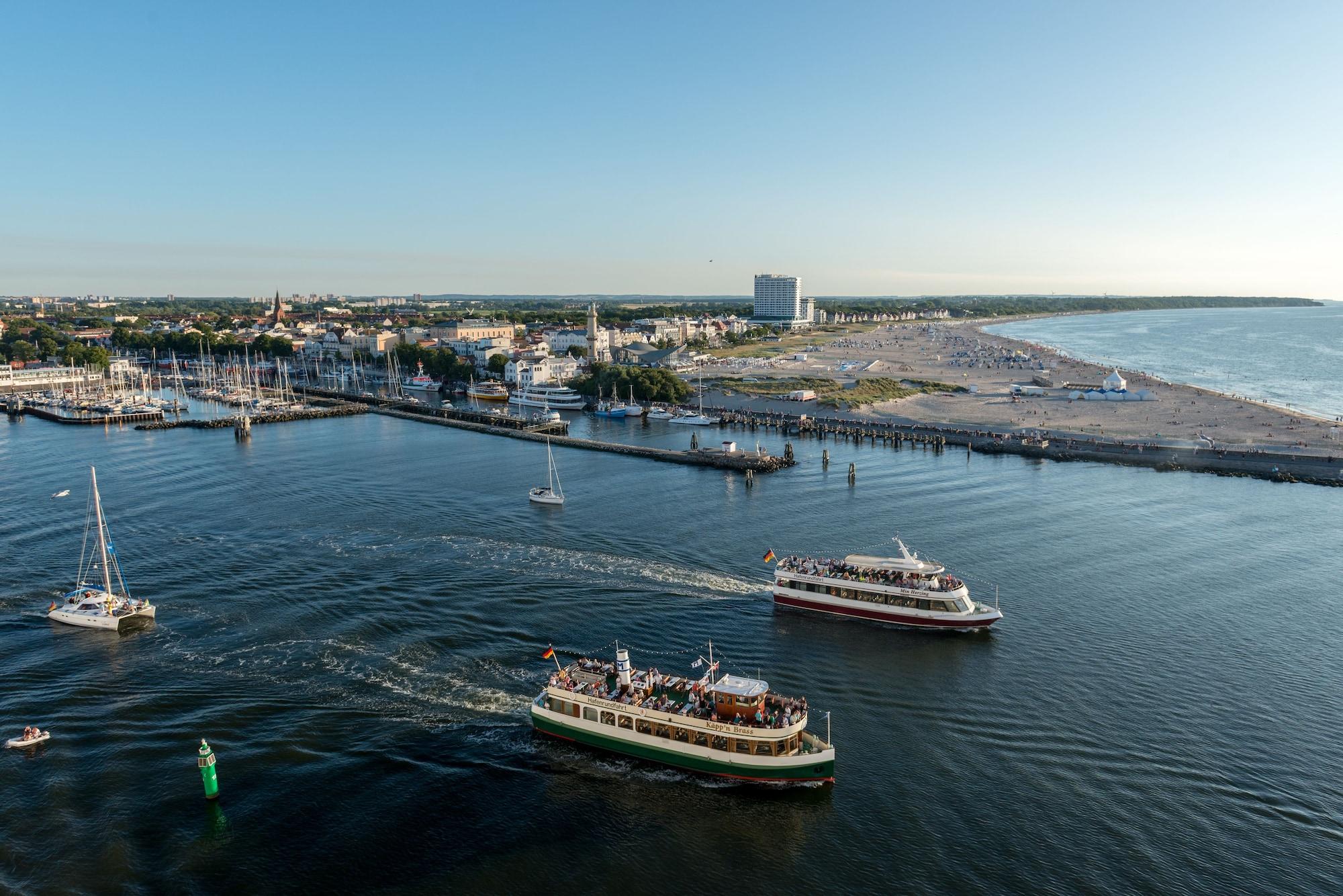 Hotel Neptun Rostock Eksteriør bilde The photo depicts a coastal scene featuring a harbor or marina. There are several boats navigating through the water, with at least two larger vessels evident, one possibly a ferry, both creating wake trails behind them. In the background, there are 