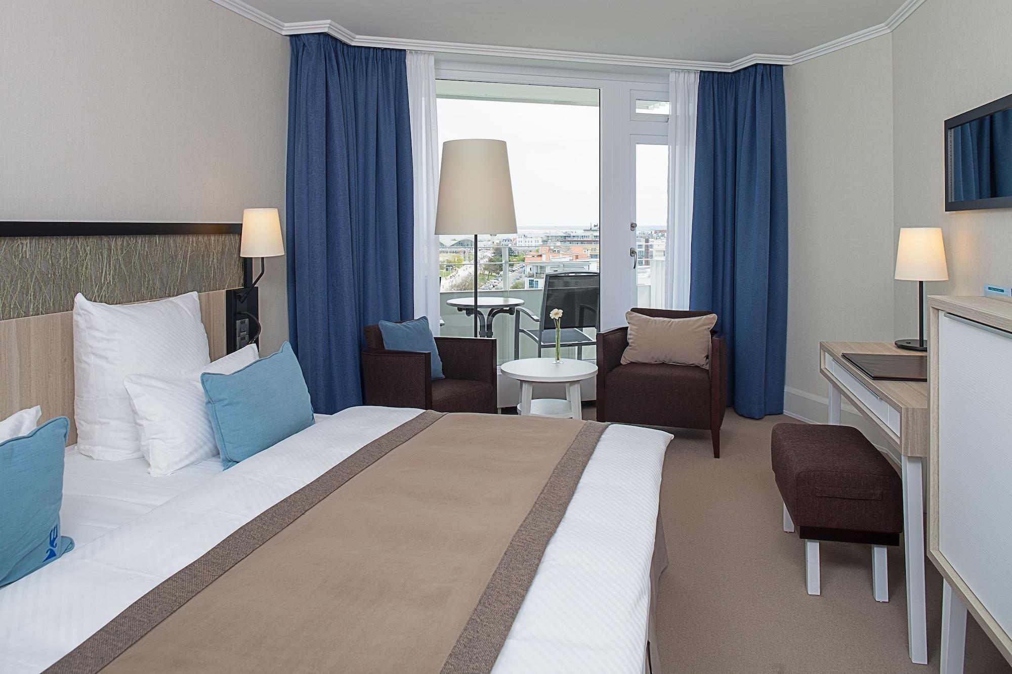 Hotel Neptun Rostock Eksteriør bilde The photo shows a modern hotel room featuring a neatly made bed with white sheets and a beige throw blanket. There are two blue pillows on the bed. The room has large windows with light blue curtains that allow natural light to enter, and a lamp is p