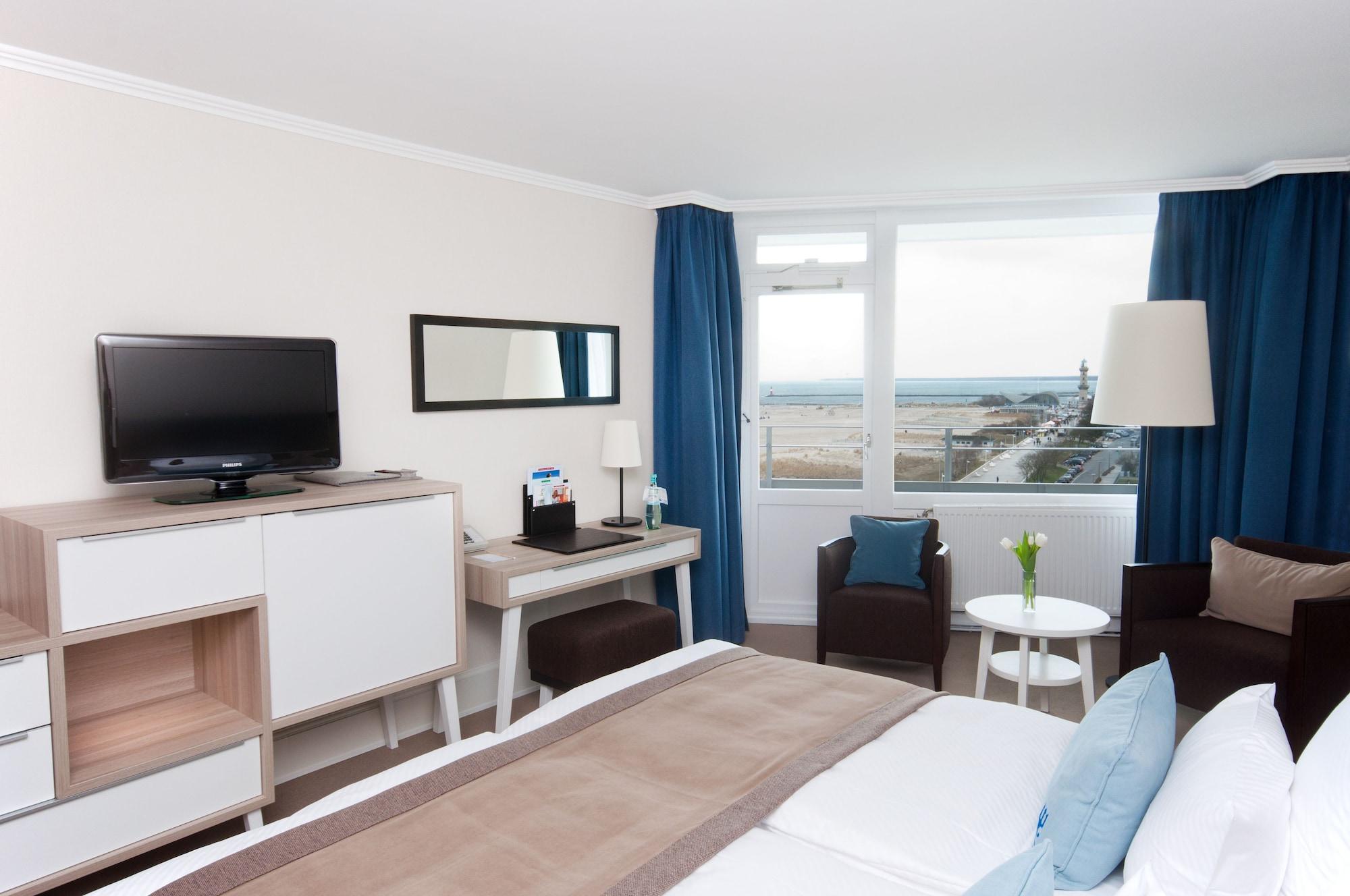 Hotel Neptun Rostock Eksteriør bilde The photo shows a modern hotel room. It features a neatly made bed with light-colored linens. Opposite the bed is a wall-mounted flat-screen TV, accompanied by a console and a mirror. There is a small desk area with a chair, and next to it, a round t