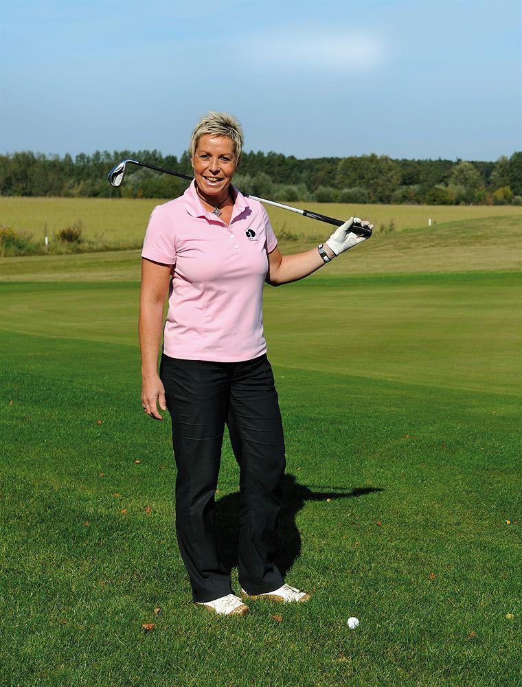 Hotel Neptun Rostock Eksteriør bilde The image shows a woman standing on a golf course, dressed in a pink polo shirt and black pants. She is holding a golf club over her shoulder with one hand and smiling at the camera. The background features a grassy area and trees, indicating a sunny