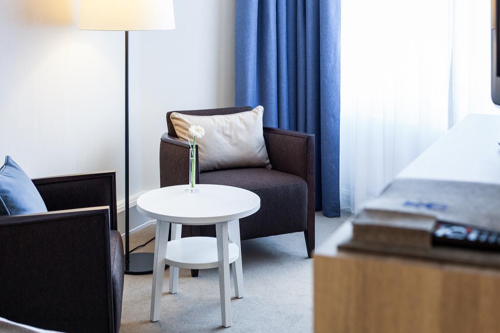 Hotel Neptun Rostock Eksteriør bilde The photo shows a cozy corner of a room featuring two armchairs—one dark brown and the other light blue. Between them is a small white table with a flower in a vase on top. There's a floor lamp next to the brown chair, and a set of neutral-colored cu