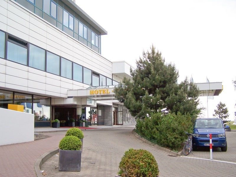 Hotel Neptun Rostock Eksteriør bilde The photo shows the exterior of a hotel building. The structure features a modern design with large windows and a sign prominently displaying the word "HOTEL." In front of the hotel, there are landscaped areas with plants and shrubs. A blue car is pa
