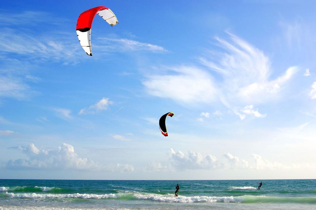 Hotel Neptun Rostock Eksteriør bilde The photo shows a beach scene with two kite surfers riding the waves. They are being pulled by colorful kites—a red and black one, and a yellow and black one—soaring high in the blue sky. The ocean appears calm with gentle waves, and the sky is partl