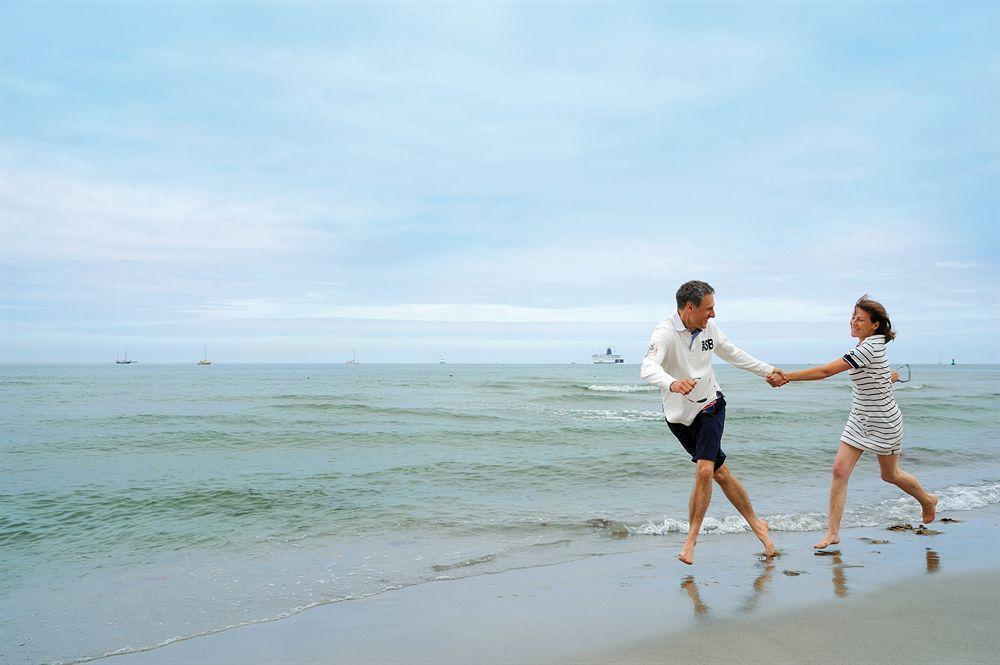 Hotel Neptun Rostock Eksteriør bilde The photo shows a couple running along the beach, holding hands. The scene is set by the ocean, with gentle waves lapping at the shore. The sky is partly cloudy, creating a calm and serene atmosphere. The man is wearing a light-colored long-sleeve sh