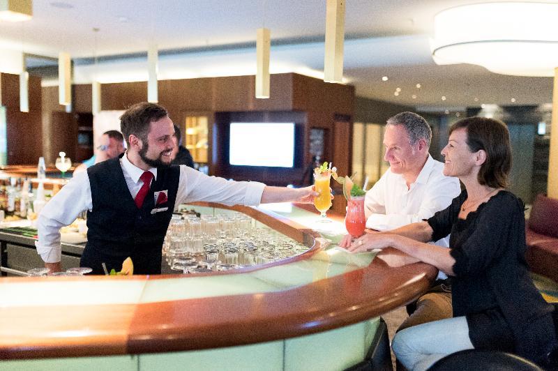 Hotel Neptun Rostock Eksteriør bilde The photo shows a bartender serving colorful cocktails to two customers at a bar. The bartender, dressed in a black vest and white shirt, is handing over drinks to a man and a woman who are seated at the bar. The setting appears to be a modern and up