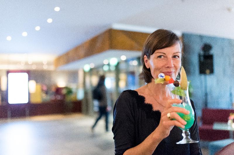 Hotel Neptun Rostock Eksteriør bilde The photo shows a woman holding a colorful tropical drink in a stylish glass. The drink is bright green and garnished with fruits and colorful decorations like a small umbrella and a straw. The setting appears to be an upscale venue, with soft lighti