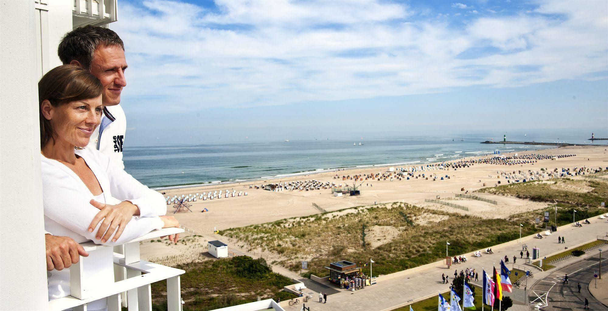 Hotel Neptun Rostock Eksteriør bilde The photo shows a scenic view of a beach with a wide stretch of sand and the ocean in the background. There are numerous beach umbrellas arranged in rows on the sand, suggesting that people are enjoying a day at the beach. In the foreground, a person