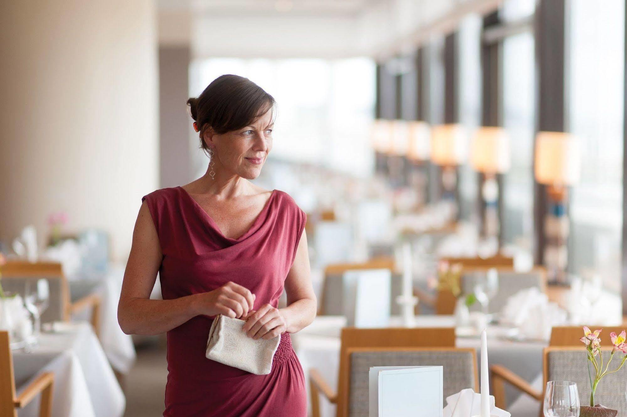 Hotel Neptun Rostock Eksteriør bilde The photo depicts a woman standing in a restaurant setting. She is dressed in a stylish red dress and holding a small purse. The restaurant has an elegant atmosphere, with tables set for dining, featuring white tablecloths and neatly arranged tablewa