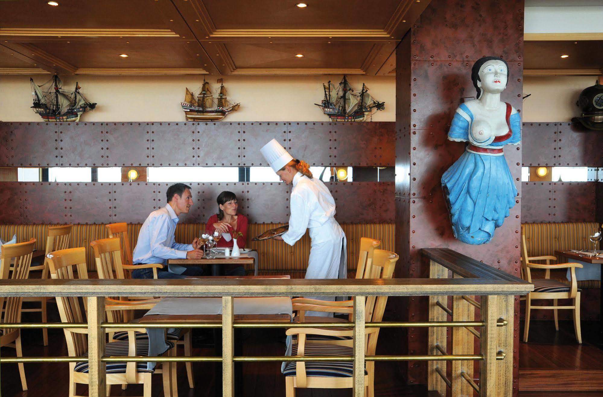 Hotel Neptun Rostock Eksteriør bilde The photo depicts a restaurant setting featuring a couple at a table with a waiter or waitress. The couple appears to be engaged in conversation while being served. The staff member is dressed in a chef’s uniform, complete with a white hat. There are