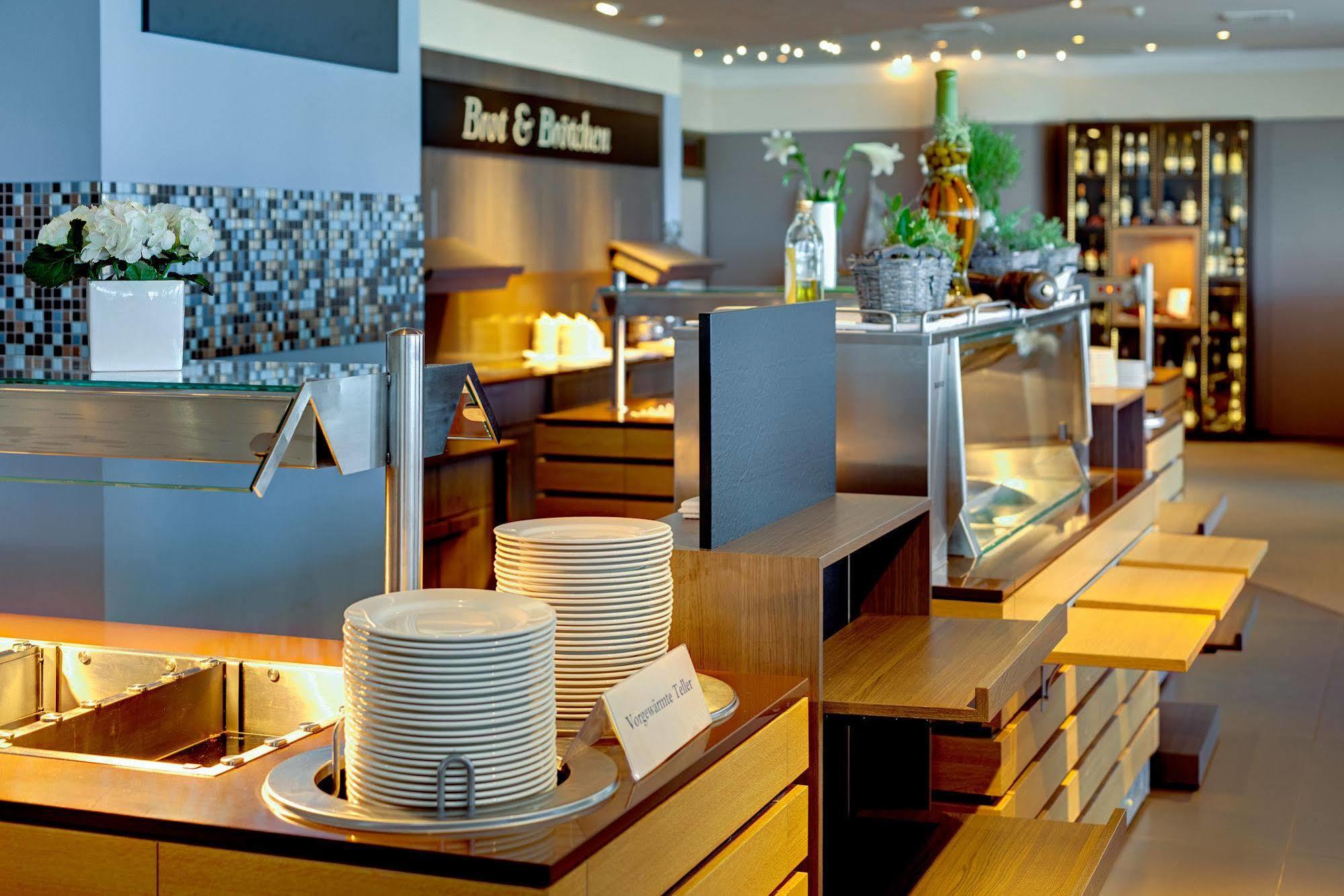 Hotel Neptun Rostock Eksteriør bilde The photo shows a modern dining area, likely a buffet restaurant or a dining hall. There are neatly arranged stacks of white plates on a wooden counter, and various food displays behind glass. The decor appears sleek and contemporary, with well-lit a