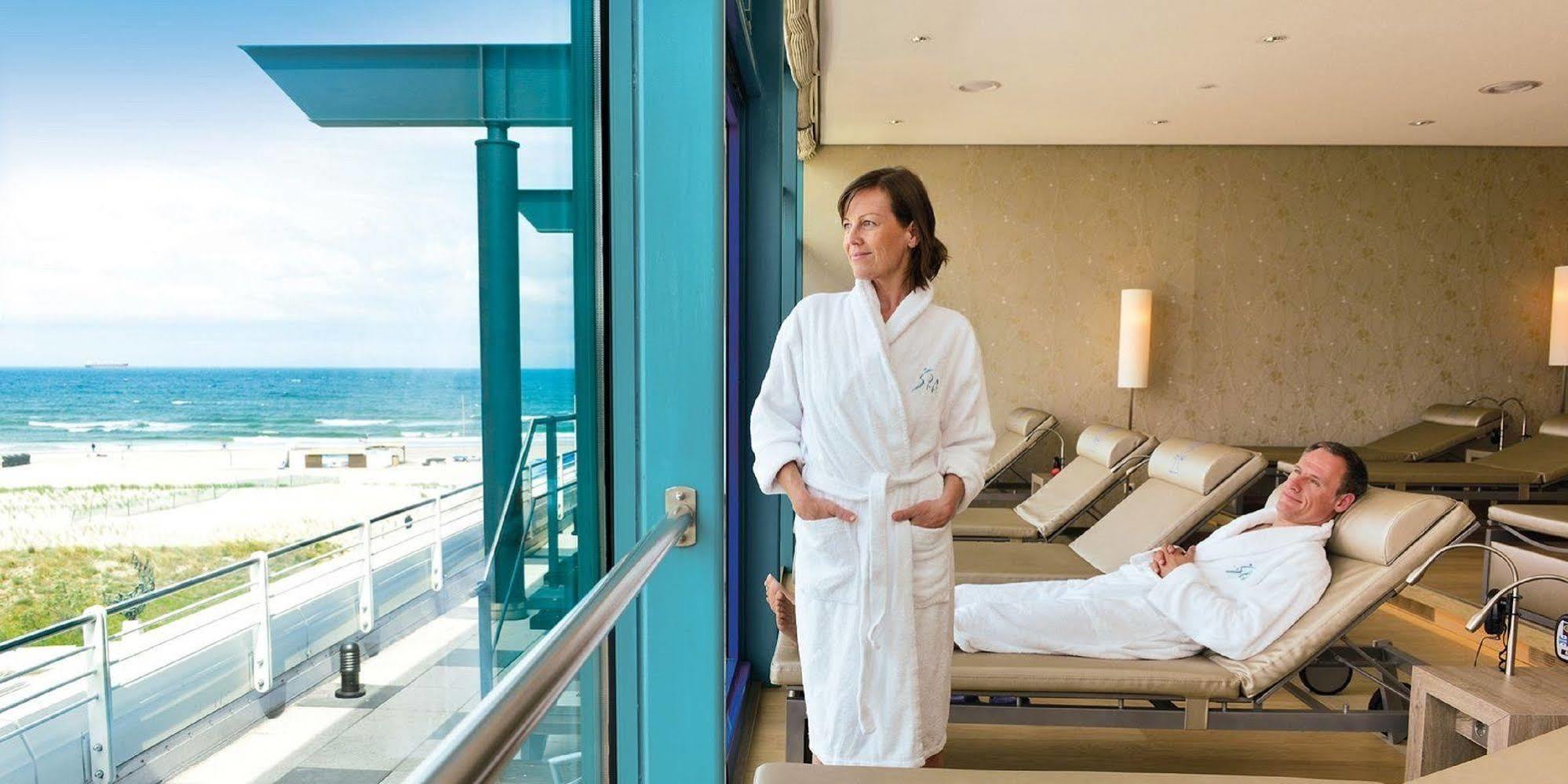 Hotel Neptun Rostock Eksteriør bilde The photo shows a woman in a white bathrobe standing near a large window with a view of the ocean. She appears to be relaxed and contemplative, looking out at the water. In the background, there are lounge chairs, and another person is lying down, su