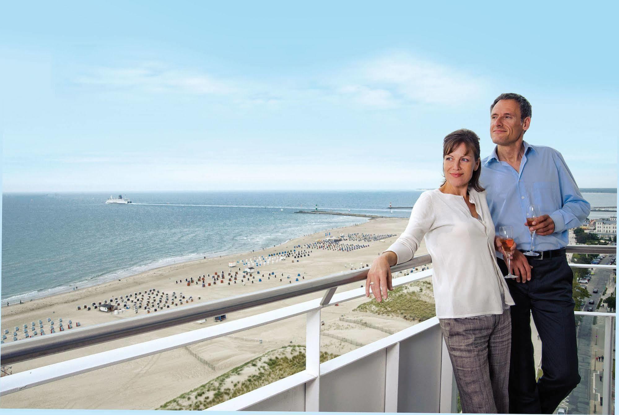 Hotel Neptun Rostock Eksteriør bilde The photo shows a couple standing on a balcony overlooking a beach. They appear to be enjoying a scenic view of the ocean and coastline. The beach has some umbrellas or beach chairs arranged in rows, and the sky is clear with a bright blue tone. The 