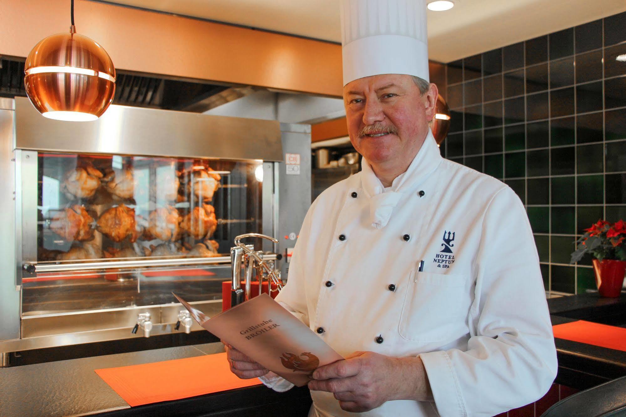 Hotel Neptun Rostock Eksteriør bilde The photo shows a chef standing in a restaurant kitchen. He is wearing a white chef's uniform and a tall chef hat. The chef is holding a menu or a dish list and smiling. Behind him, there is a large oven or grill with roasted items visible, likely co