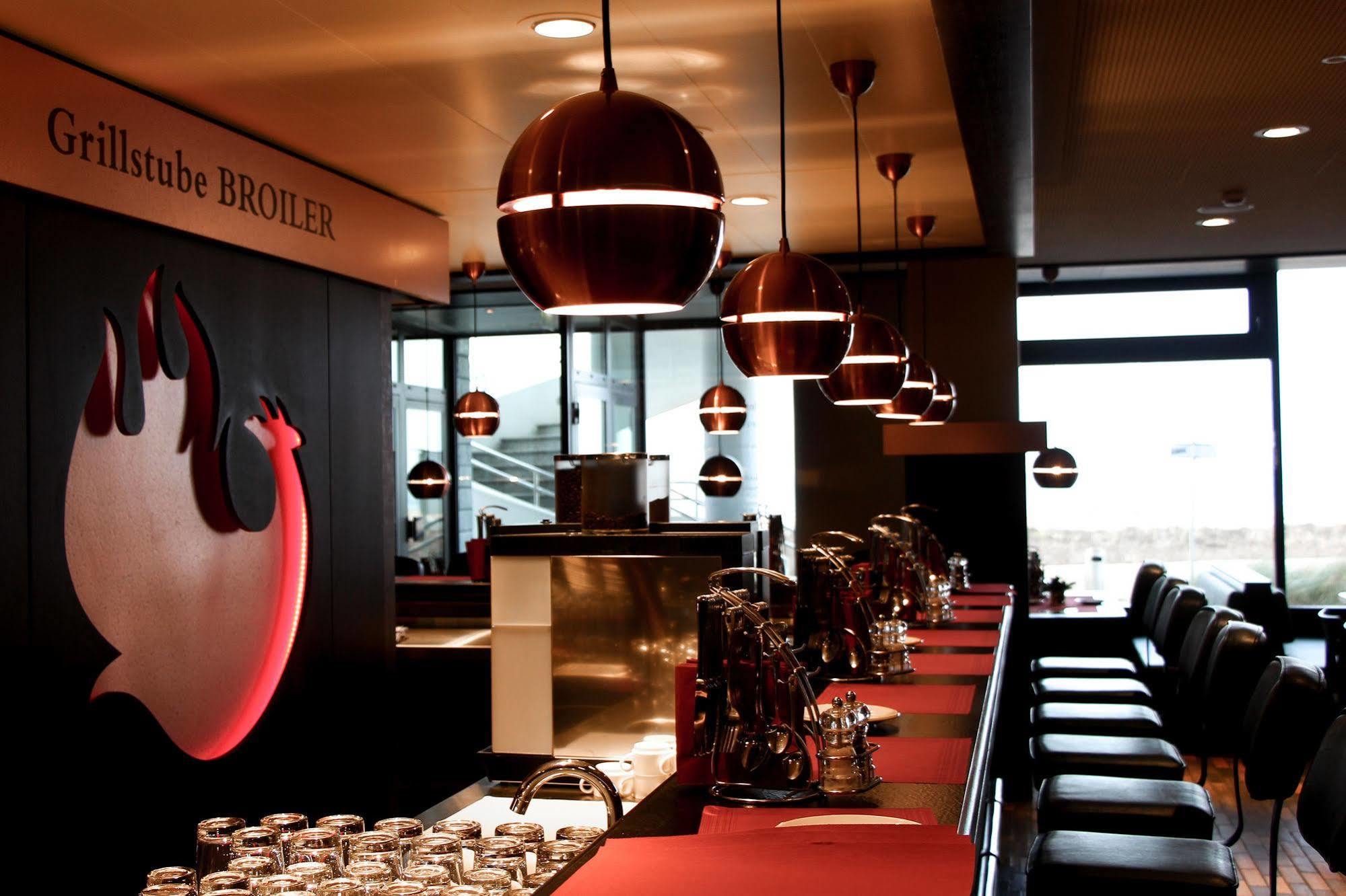 Hotel Neptun Rostock Eksteriør bilde The photo depicts the interior of a modern dining establishment, likely a restaurant or grill. The design features a long bar or counter with a red surface, complemented by black seating. Pendant lights with a circular design hang from the ceiling, p