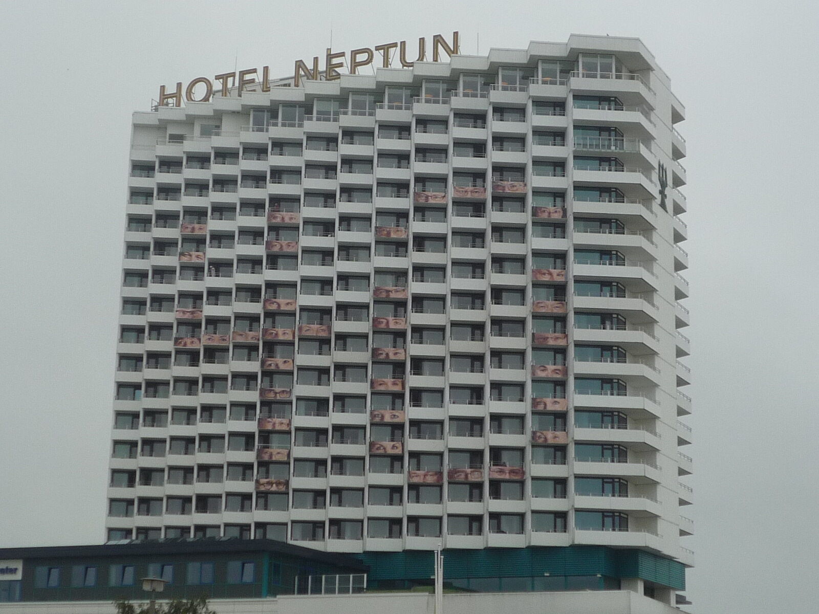 Hotel Neptun Rostock Eksteriør bilde The photo shows a tall, modern hotel building named "Hotel Neptun." It has a white facade with multiple rectangular windows and balconies. The top of the building features the hotel name prominently displayed. The sky appears overcast, indicating clo